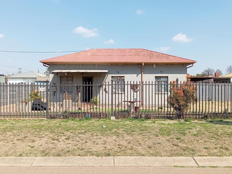 Front elevation of a single-story detached house featuring a corrugated metal roof, brick exterior, and secure perimeter steel fencing.
