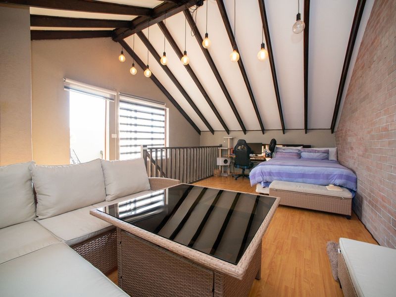 Spacious loft-style bedroom featuring exposed timber ceiling beams, a rustic brick feature wall, and warm timber laminate flooring with an open-plan layout.