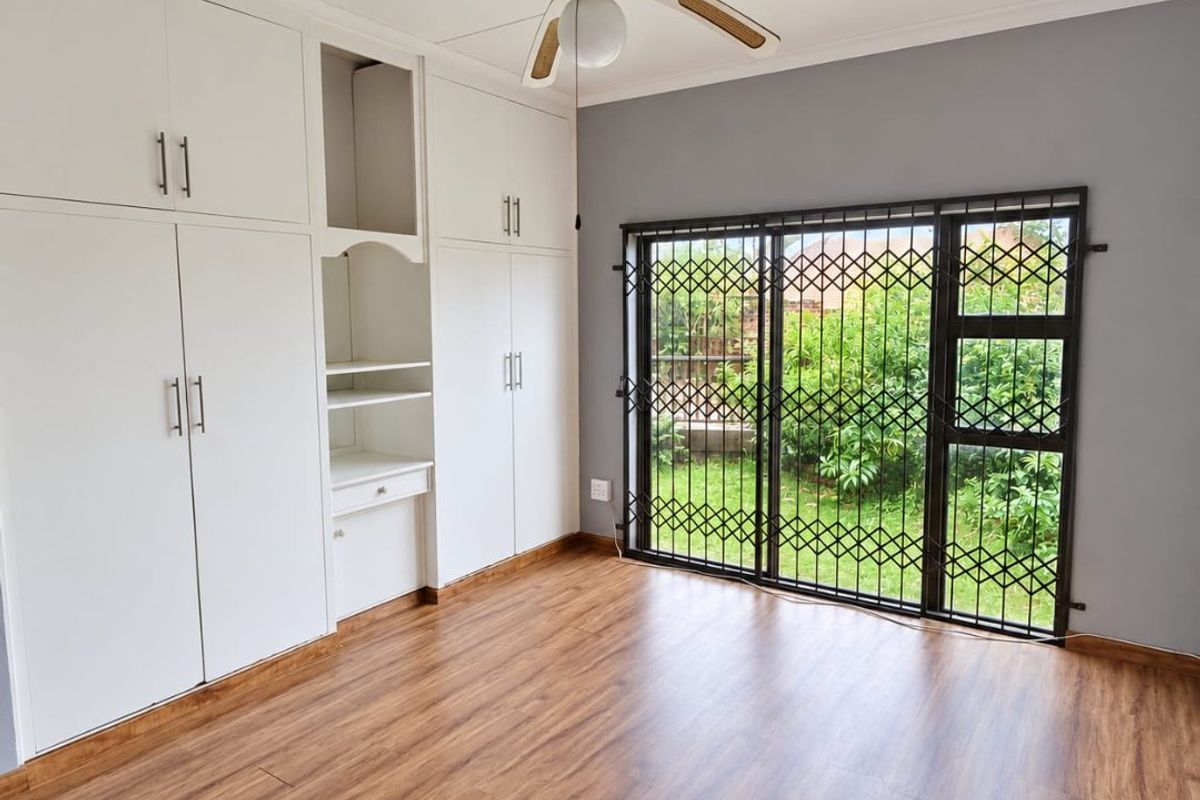 Spacious bedroom featuring wood-look laminate flooring, extensive built-in cabinetry, a ceiling fan, and sliding glass doors with security gates leading to the garden.