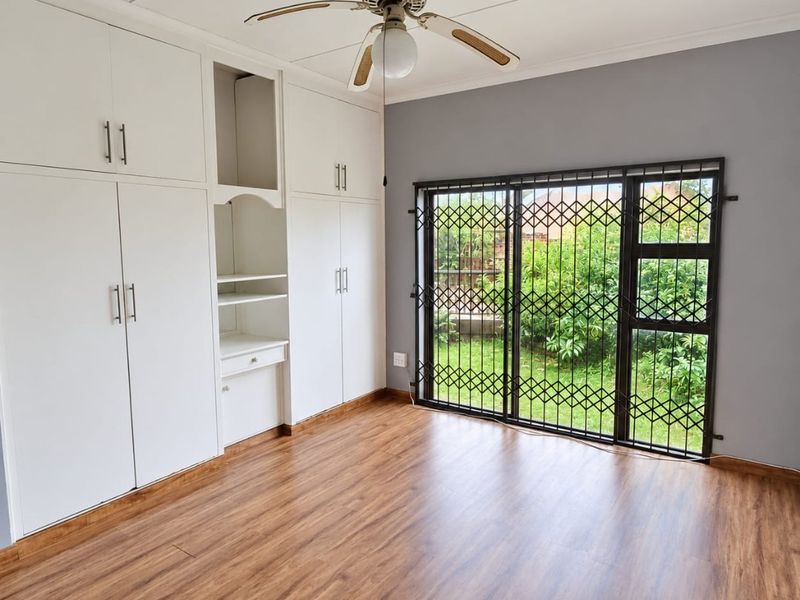 Spacious bedroom featuring wood-look laminate flooring, extensive built-in cabinetry, a ceiling fan, and sliding glass doors with security gates leading to the garden.