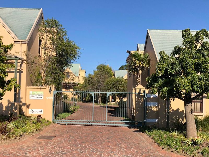 Front entrance view of a commercial property complex featuring a secure metal gate, brick paved driveway, and well-maintained stucco buildings with pitched roofs.