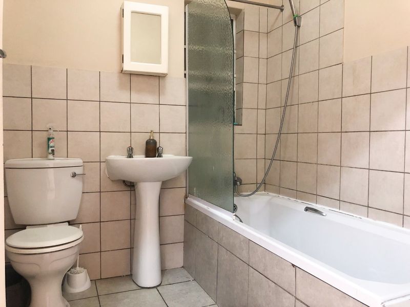 A functional bathroom featuring a pedestal basin, toilet, and a bathtub with a glass shower screen, finished with neutral ceramic wall and floor tiles.