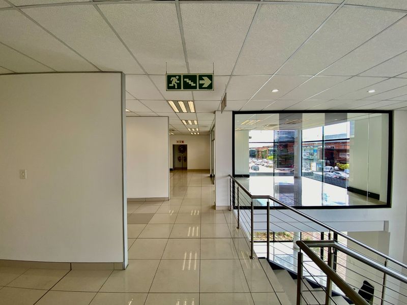 A bright, modern industrial hallway featuring tiled floors, a suspended ceiling with recessed lighting, and a glass partition overlooking the lower level.