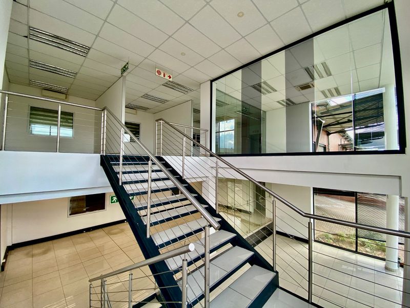Modern industrial office interior featuring a central staircase with stainless steel balustrades, glass partitioning, and clean tiled flooring throughout the open-plan space.