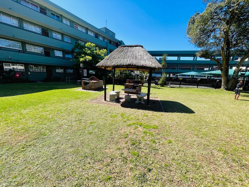 Communal garden area featuring a thatched lapa with a built-in brick braai and paved seating space, set within the apartment complex grounds.