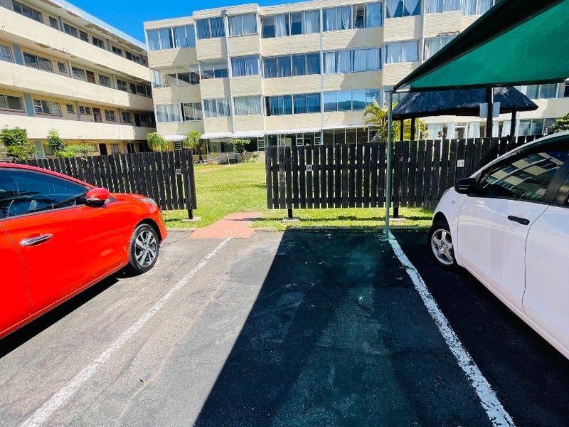 View of the apartment complex rear exterior featuring designated parking bays, a shade cloth carport, and a communal garden area enclosed by timber fencing.