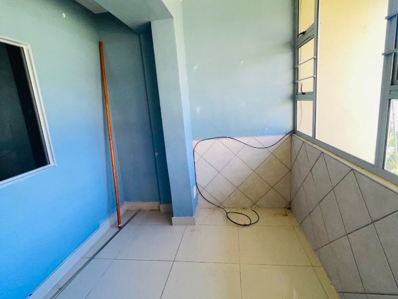 An enclosed balcony or sunroom featuring tiled flooring, tiled wall accents, and aluminium-framed windows, currently vacant and requiring minor cosmetic updates.