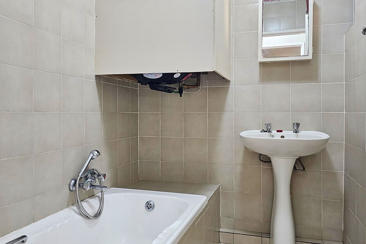 A functional bathroom featuring a pedestal basin, built-in bathtub, and wall-mounted geyser with tiled walls throughout.