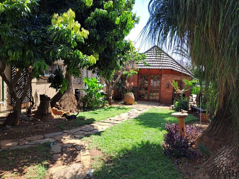 A lush, established garden featuring a stone pathway leading to a charming brick outbuilding with a tiled roof and french doors.