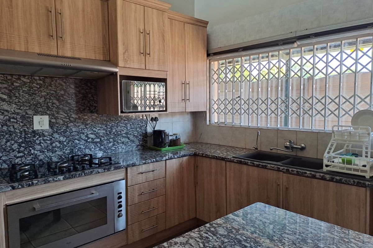 A well-maintained kitchen featuring wooden cabinetry, granite countertops, a gas hob, built-in oven, and a double basin sink with security bars on the window.