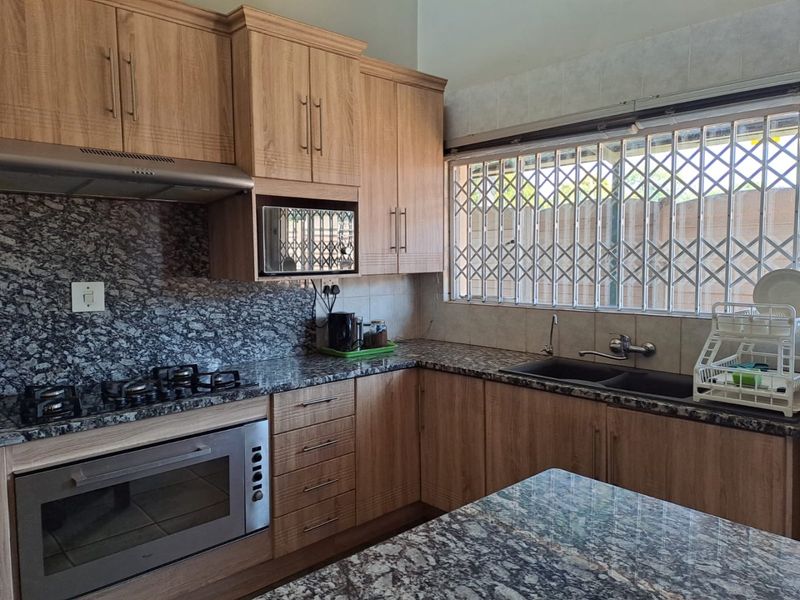 A well-maintained kitchen featuring wooden cabinetry, granite countertops, a gas hob, built-in oven, and a double basin sink with security bars on the window.