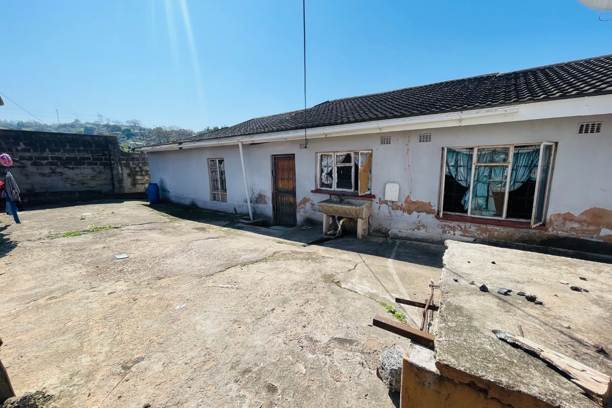 Rear exterior view of the property featuring a concrete patio, external wash basin, and a boundary wall, showing significant signs of wear and maintenance needs.
