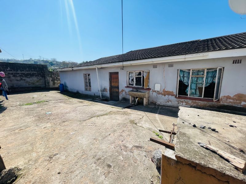 Rear exterior view of the property featuring a concrete patio, external wash basin, and a boundary wall, showing significant signs of wear and maintenance needs.
