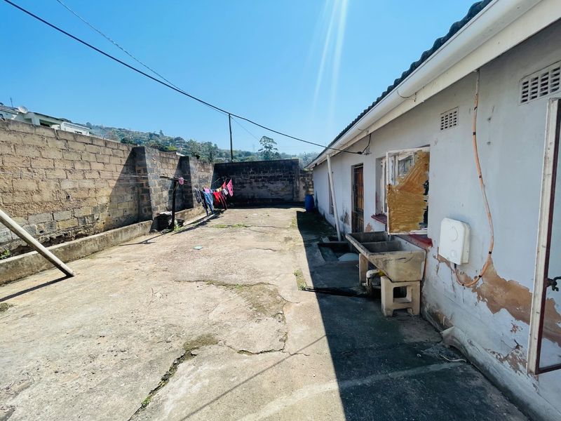 Rear exterior area featuring concrete paving, a perimeter boundary wall, and an external utility sink, showing signs of wear and maintenance requirements.