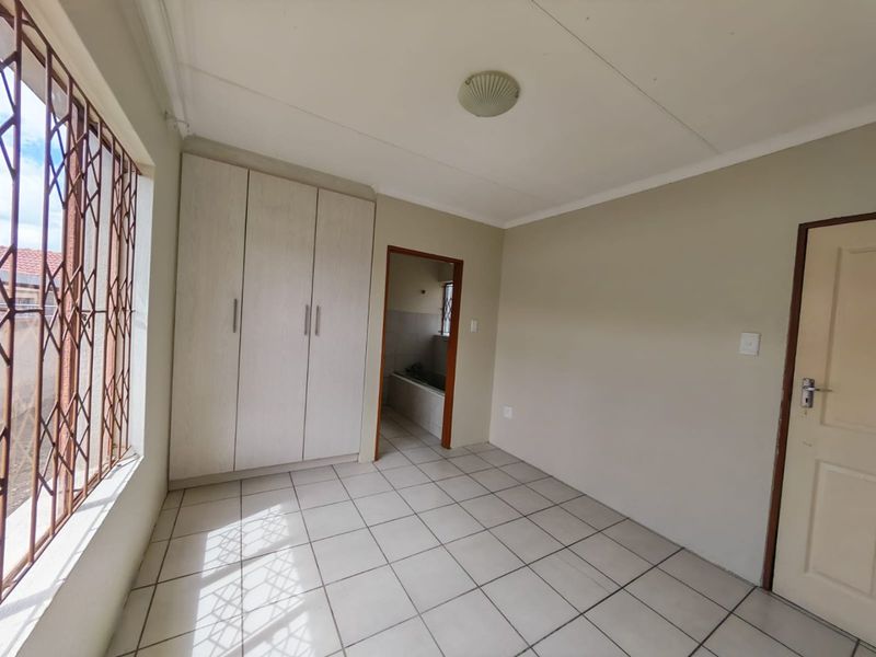 A clean, empty bedroom featuring tiled flooring, built-in cupboards, and security bars on the window, with an adjoining bathroom visible through the doorway.