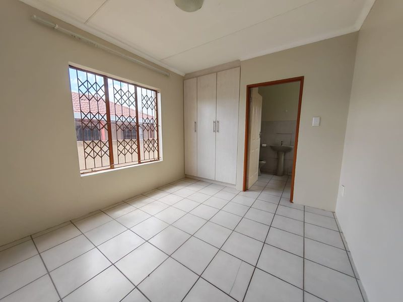 A clean, empty bedroom featuring tiled flooring, built-in cupboards, and security bars on the window, with an adjoining en-suite bathroom.