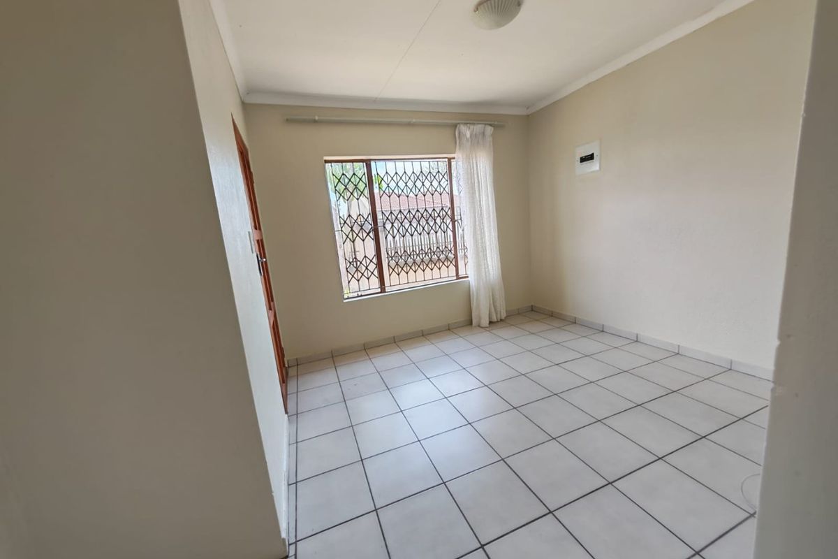 A clean, empty bedroom featuring neutral tiled flooring, a window with security bars, and a standard electrical distribution board.