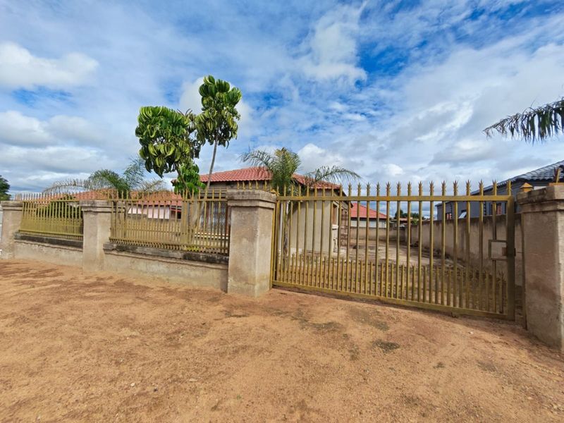 Front view of the property featuring a secure metal perimeter fence, concrete pillars, and a gated entrance with a tiled roof visible in the background.