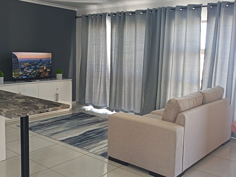 A well-maintained living room featuring neutral tiled flooring, a dark accent wall, and large windows dressed with floor-to-ceiling curtains.