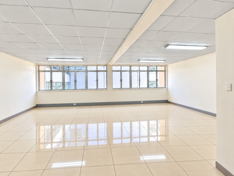 A bright, vacant commercial office space featuring polished tiled flooring, a suspended ceiling with integrated lighting, and large windows providing ample natural light.