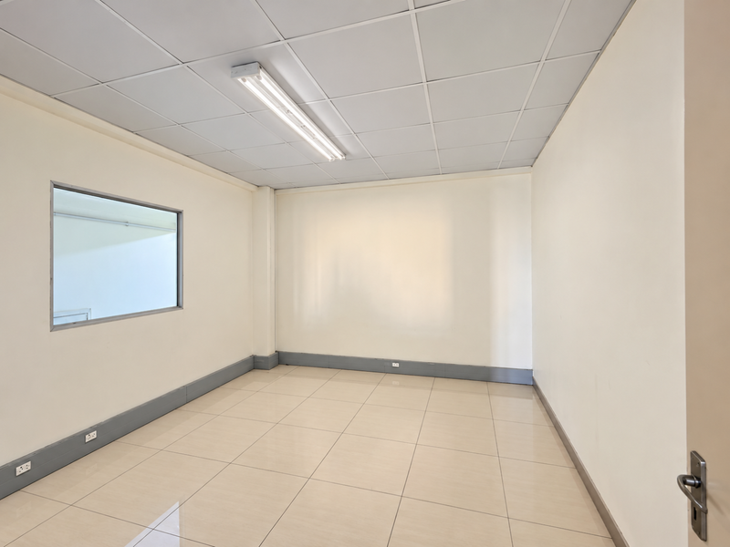 A clean, empty office space featuring tiled flooring, a suspended ceiling with integrated lighting, and an internal window providing natural light.