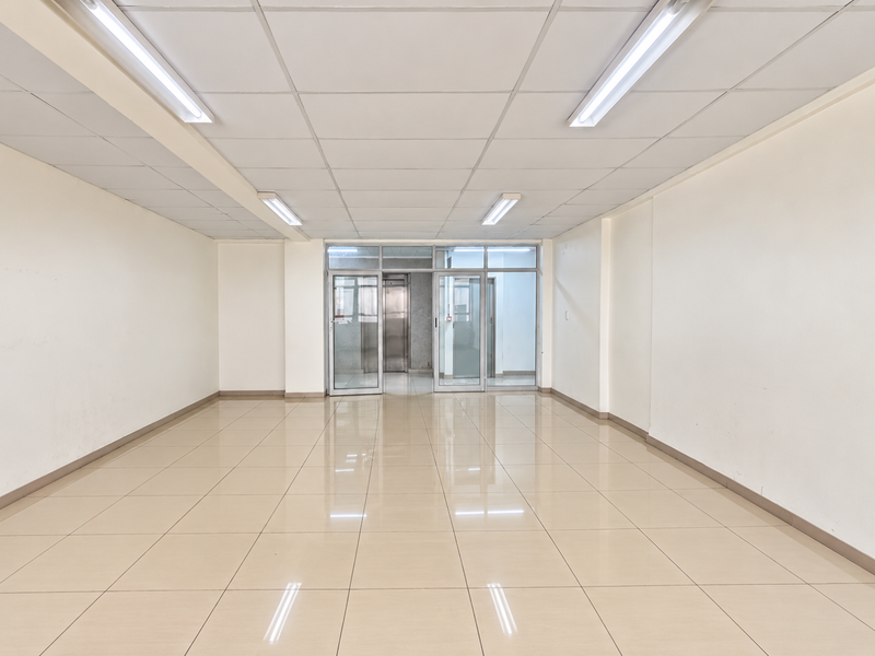A clean, vacant commercial office space featuring polished ceramic tile flooring, a suspended ceiling with integrated lighting, and glass entry doors with elevator access.