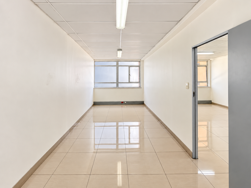 A bright, empty commercial hallway featuring tiled flooring, suspended ceilings with fluorescent lighting, and clean white walls leading to an adjacent office space.
