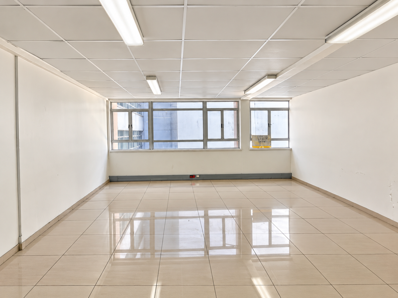 A bright, empty commercial office space featuring tiled flooring, a suspended ceiling with integrated lighting, and large windows providing natural light.