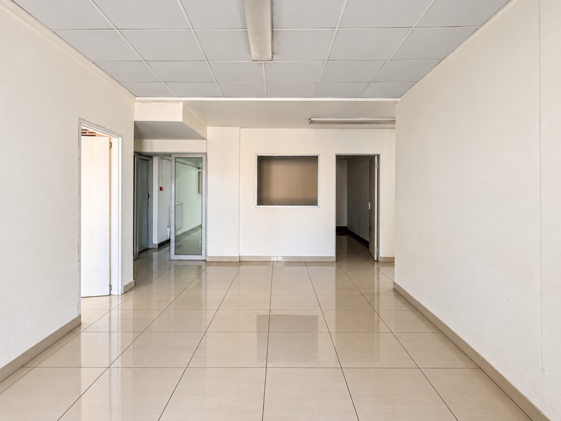 A bright, vacant commercial office space featuring neutral ceramic tile flooring, a suspended ceiling with integrated lighting, and multiple internal access points.
