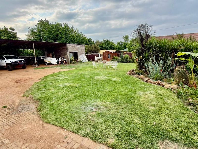 Spacious backyard featuring a large lawn, established garden beds, a paved driveway, and a covered carport area for vehicle storage.
