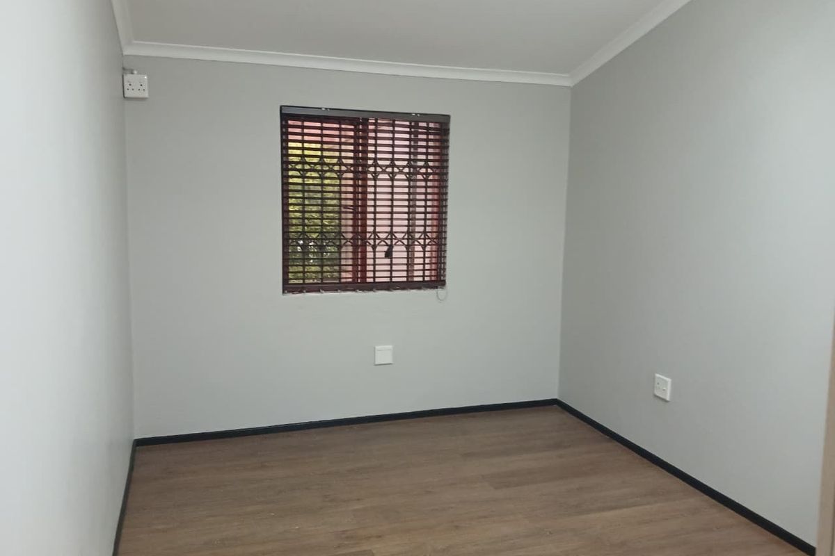 A clean, empty bedroom featuring neutral grey walls, modern laminate flooring, and a window fitted with security bars and venetian blinds.