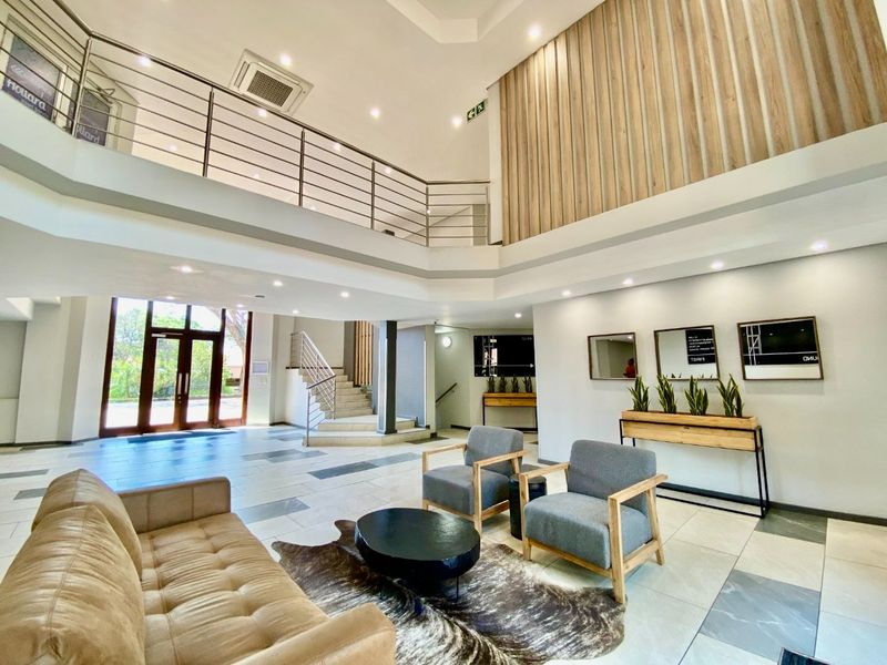 Spacious commercial entrance lobby featuring double volume ceilings, a mezzanine level with stainless steel balustrades, modern tiled flooring, and contemporary timber wall accents.