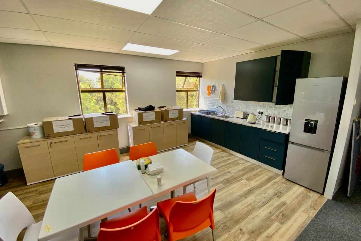 A functional commercial kitchen area featuring modern dark cabinetry, light wood-look flooring, a tiled backsplash, and ample natural light from two windows.