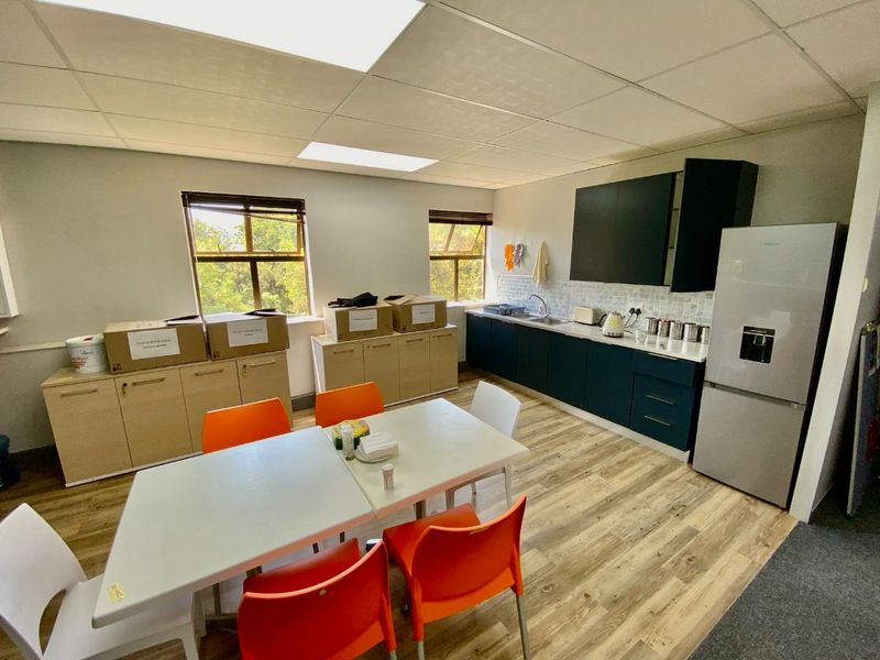 A functional commercial kitchen area featuring modern dark cabinetry, light wood-look flooring, a tiled backsplash, and ample natural light from two windows.
