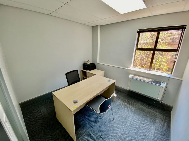 A functional commercial office space featuring carpeted flooring, a suspended ceiling, and a window with an integrated HVAC unit.