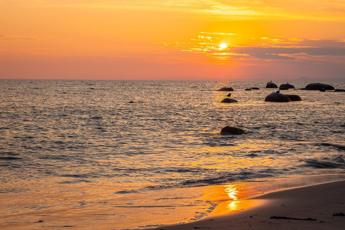 A scenic sunset view over the ocean, featuring a sandy shoreline and rocky outcrops in the water.