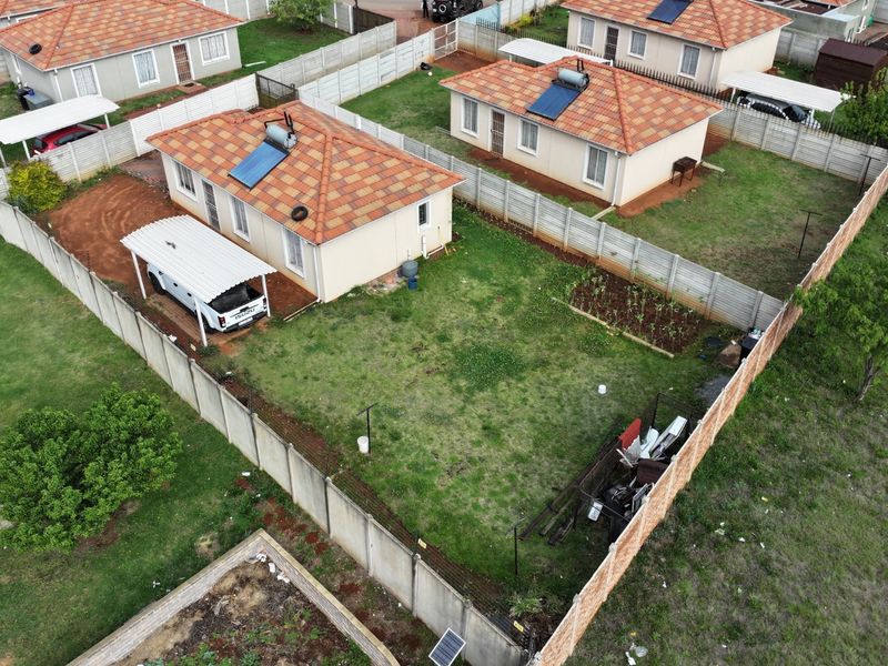 Aerial view of a residential property featuring a tiled roof house with a solar water heater, carport, and a fenced garden area.