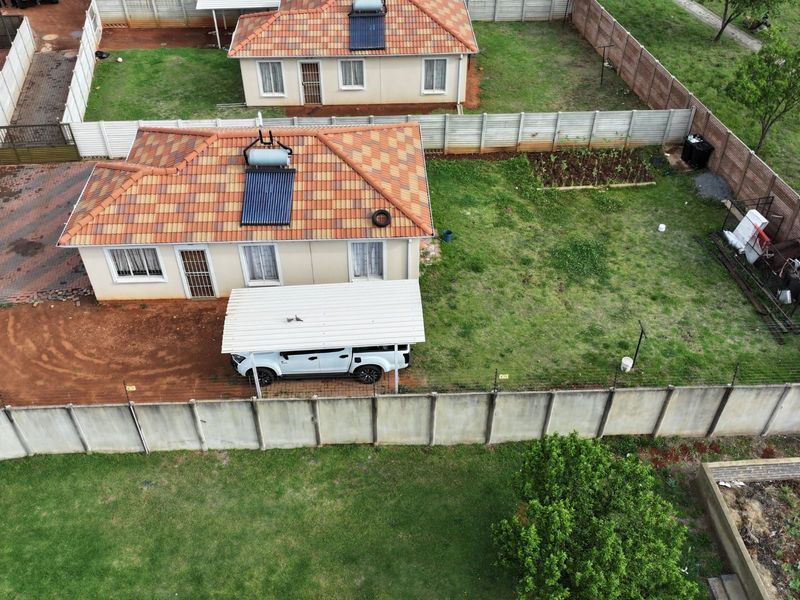 Aerial view of a residential property featuring a tiled roof house with a solar water heater, carport, and a spacious garden with boundary walling.