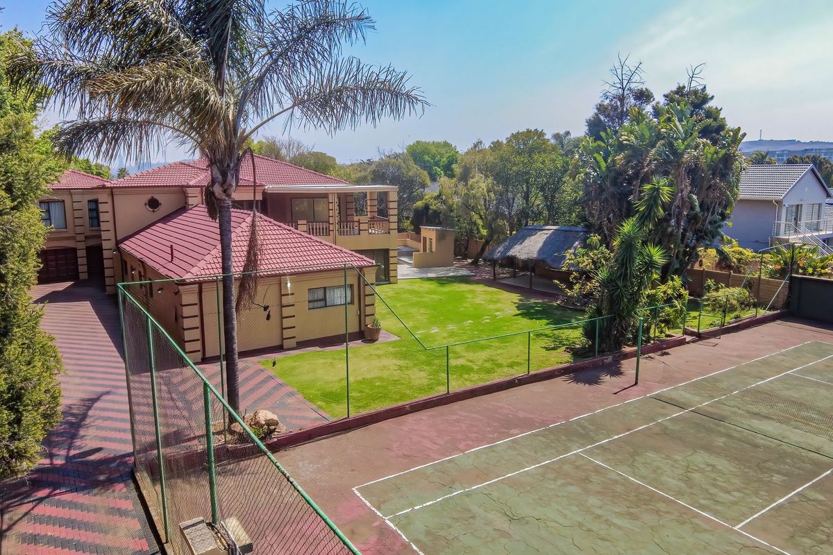 Aerial view of a residential property featuring a private tennis court, a thatched lapa, a large garden, and a paved driveway.