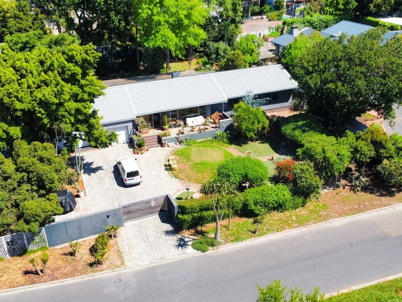 Aerial view of a residential property featuring a metal roof, paved driveway, sliding gate, and a well-maintained landscaped garden with a patio area.