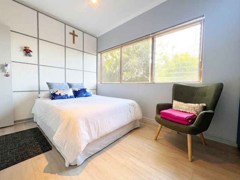 A well-lit bedroom featuring light laminate flooring, built-in wall paneling with integrated cupboards, and large windows fitted with venetian blinds.