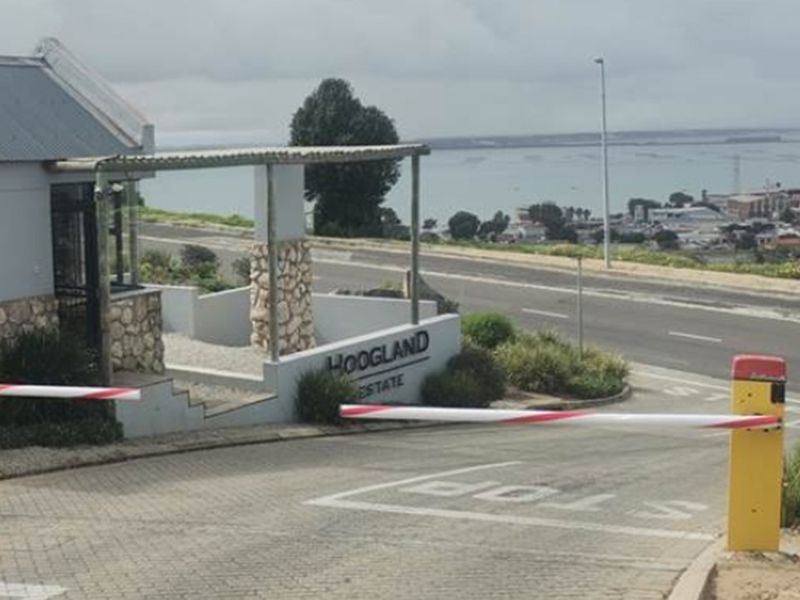 Entrance view of Hoogland Estate featuring a security boom gate, paved driveway, and stone-clad guardhouse structure overlooking the coastal landscape.