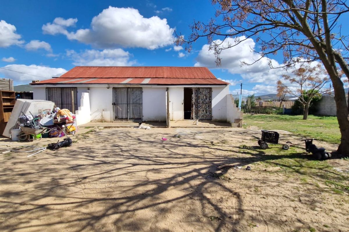 Rear view of a single-story dwelling featuring a corrugated metal roof and security gates, set on a large plot with unlandscaped grounds.