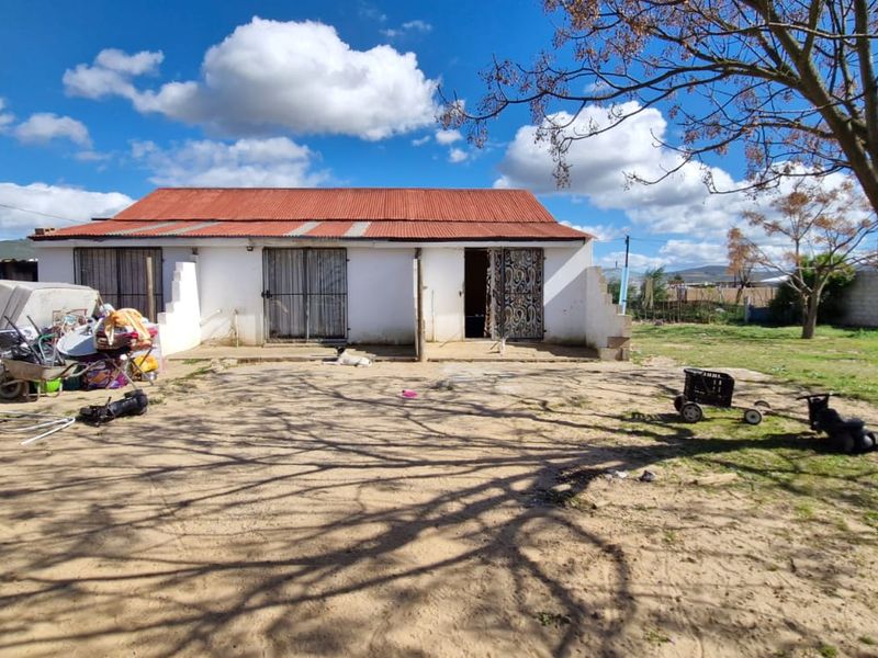 Rear view of a single-story dwelling featuring a corrugated metal roof and security gates, set on a large plot with unlandscaped grounds.