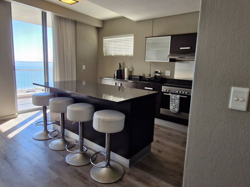 Modern open-plan kitchen and living area featuring a stone-topped island, dark cabinetry, and direct access to a balcony with ocean views.