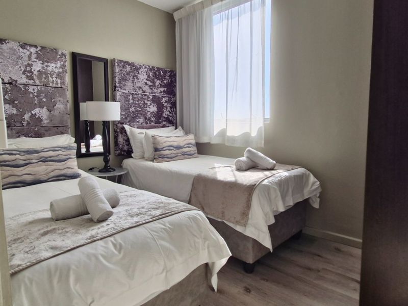 A compact bedroom featuring laminate flooring and a window with sheer curtains, currently configured with two single beds.