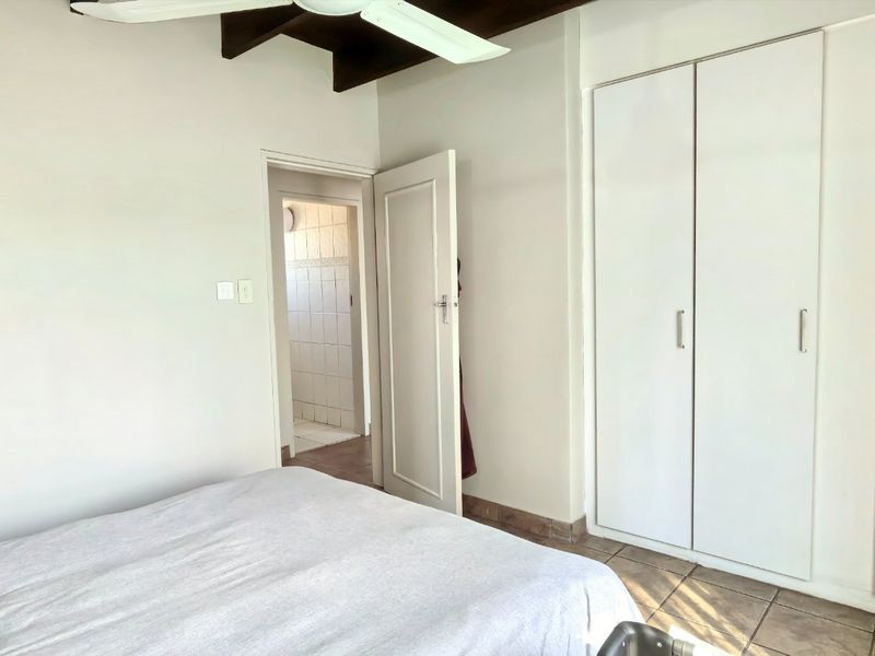 Bedroom featuring built-in cupboards, tiled flooring, and exposed timber ceiling beams with a ceiling fan, leading to an adjacent bathroom.