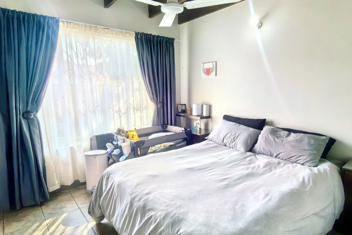 A bedroom featuring tiled flooring, exposed timber ceiling beams, and a ceiling fan, with natural light entering through a large window with security gate.