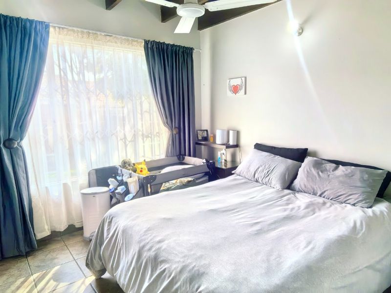 A bedroom featuring tiled flooring, exposed timber ceiling beams, and a ceiling fan, with natural light entering through a large window with security gate.