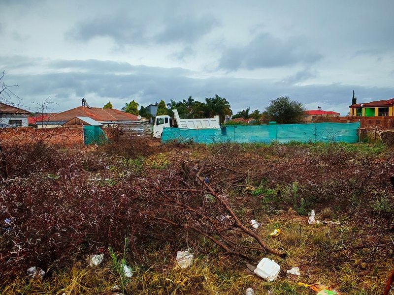 A vacant residential plot featuring a brick boundary wall and green shade cloth fencing, currently requiring clearing of vegetation and debris.